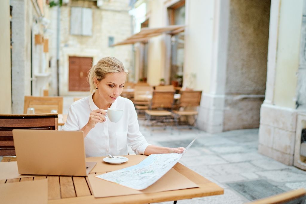 Work and travel. Pretty young woman with map sitting at table using laptop computer at old town cafe, outside.